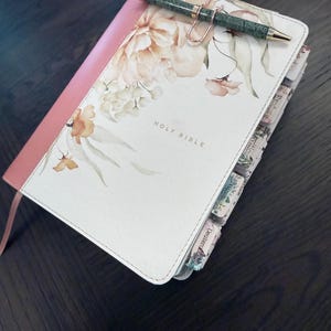 Puede incluir: Una Santa Biblia con cubierta blanca con estampado floral, lomo rosa y texto en tono dorado. El libro incluye pesta&ntilde;as de &iacute;ndice en el lateral y un bol&iacute;grafo con estampado verde sujeto por un clip de metal en oro rosa sobre la portada.