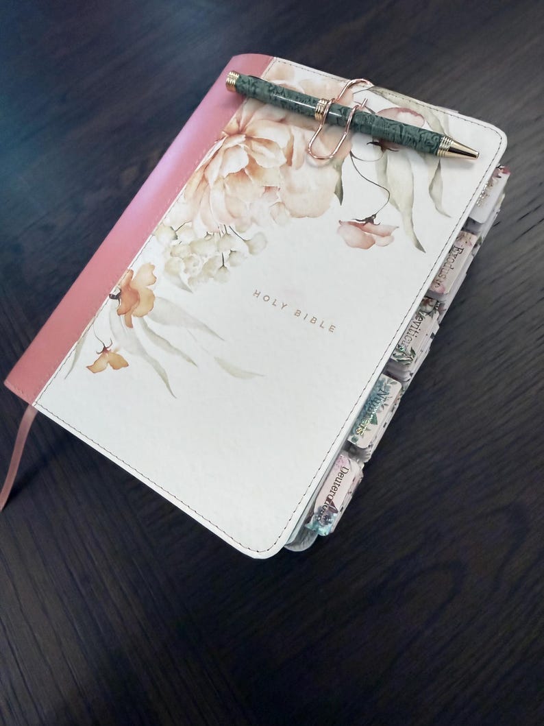 Puede incluir: Una Santa Biblia con cubierta blanca con estampado floral, lomo rosa y texto en tono dorado. El libro incluye pesta&ntilde;as de &iacute;ndice en el lateral y un bol&iacute;grafo con estampado verde sujeto por un clip de metal en oro rosa sobre la portada.