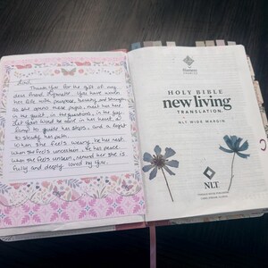 Puede incluir: Una Santa Biblia New Living Translation abierta con amplios m&aacute;rgenes. La p&aacute;gina izquierda presenta una nota escrita a mano sobre papel decorativo con patrones de flores y mariposas. La p&aacute;gina derecha muestra texto impreso y dos flores azules prensadas.