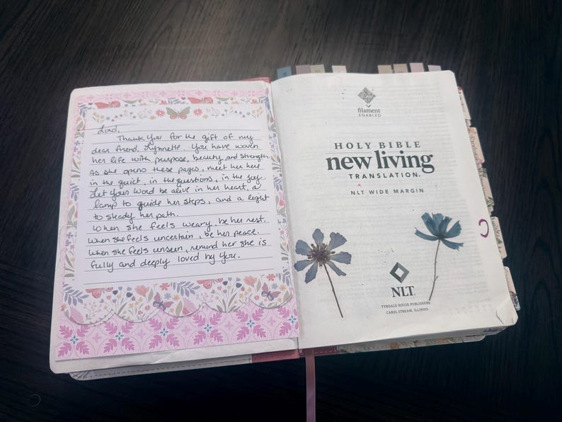 Puede incluir: Una Santa Biblia New Living Translation abierta con amplios m&aacute;rgenes. La p&aacute;gina izquierda presenta una nota escrita a mano sobre papel decorativo con patrones de flores y mariposas. La p&aacute;gina derecha muestra texto impreso y dos flores azules prensadas.
