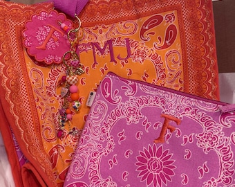 Puede incluir: Un bolso tote naranja y rojo con estampado de bandana, las letras bordadas FMJ y una etiqueta circular rosa con la letra F, acompañado de un estuche con cremallera a juego en rosa y blanco con estampado de bandana y la letra F bordada.