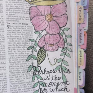 Puede incluir: Una p&aacute;gina de una Biblia con una ilustraci&oacute;n dibujada a mano de una corona dorada sobre dos flores rosas con hojas verdes. El texto "Perhaps this is the moment for which you have been created" est&aacute; escrito en tinta negra.