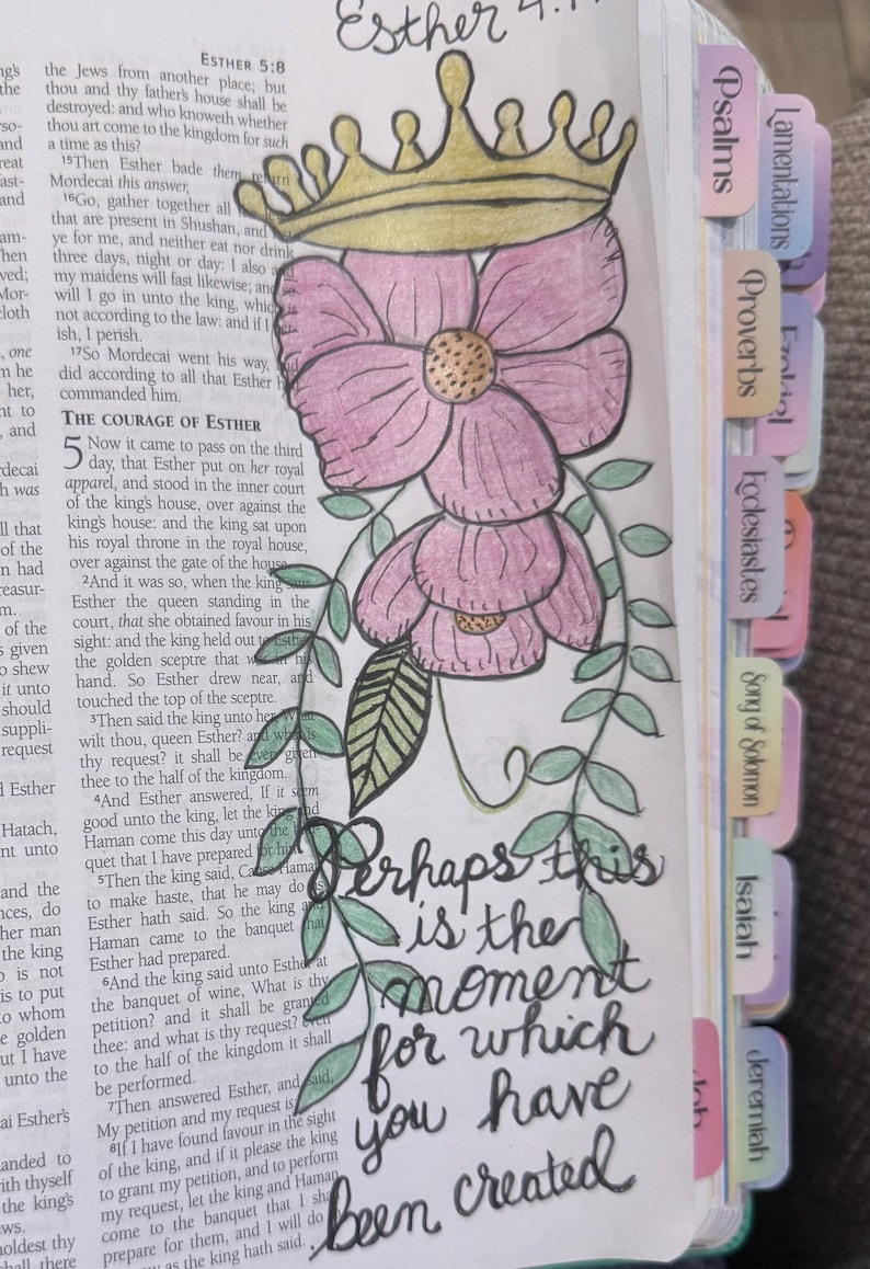 Puede incluir: Una p&aacute;gina de una Biblia con una ilustraci&oacute;n dibujada a mano de una corona dorada sobre dos flores rosas con hojas verdes. El texto "Perhaps this is the moment for which you have been created" est&aacute; escrito en tinta negra.