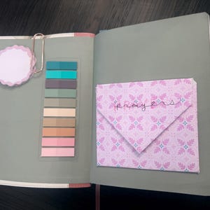 Puede incluir: Un cuaderno abierto de color verde salvia que incluye una nota adhesiva de borde festoneado sujeta por un clip de color oro rosa, una tira vertical de diez marcadores adhesivos de colores y un sobre rosa estampado con la palabra prayers.