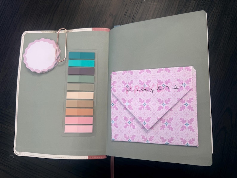 Puede incluir: Un cuaderno abierto de color verde salvia que incluye una nota adhesiva de borde festoneado sujeta por un clip de color oro rosa, una tira vertical de diez marcadores adhesivos de colores y un sobre rosa estampado con la palabra prayers.