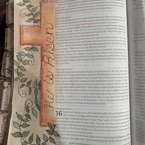 Puede incluir: Una p&aacute;gina de una Biblia con una cruz dibujada a mano en acuarela naranja y marr&oacute;n, decorada con enredaderas de hojas verdes. El texto "He is Risen" est&aacute; escrito verticalmente en tinta negra a lo largo de la cruz. Contiene texto impreso.