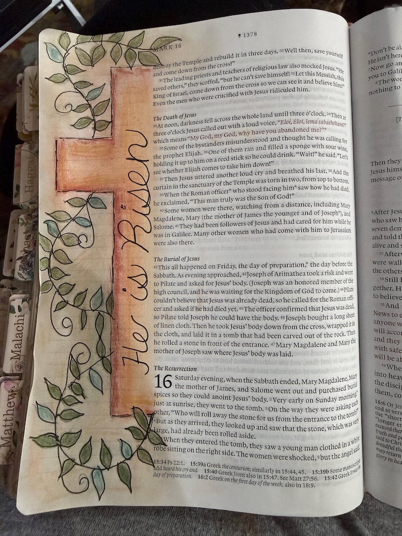 Puede incluir: Una p&aacute;gina de una Biblia con una cruz dibujada a mano en acuarela naranja y marr&oacute;n, decorada con enredaderas de hojas verdes. El texto "He is Risen" est&aacute; escrito verticalmente en tinta negra a lo largo de la cruz. Contiene texto impreso.