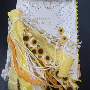 Puede incluir: Una Santa Biblia blanca con cubierta floral, adornada con un gran grupo de cintas amarillas, tiras de tela con estampado de girasoles y cuentas blancas. Un clip de papel de oro rosa sujeta un bol&iacute;grafo con estampado floral sobre el libro.
