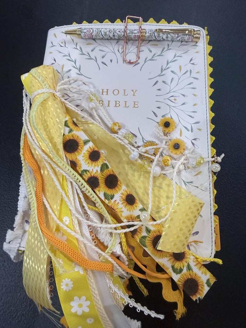 Puede incluir: Una Santa Biblia blanca con cubierta floral, adornada con un gran grupo de cintas amarillas, tiras de tela con estampado de girasoles y cuentas blancas. Un clip de papel de oro rosa sujeta un bol&iacute;grafo con estampado floral sobre el libro.