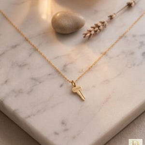 May include: A gold necklace featuring a key pendant engraved with the name "Sophia". The necklace is set against a white marble surface, with a small stone and dried flowers in the background. A delicate piece of jewellery.