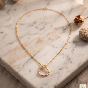 May include: A gold-colored necklace with a pendant shaped like a stylized horseshoe. The necklace chain is delicate and the pendant is a smooth, solid form. The jewelry is displayed on a marble surface.