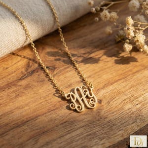 May include: A gold-colored monogram necklace with a delicate chain. The monogram features an ornate, intertwined design. The necklace is displayed on a wooden surface, with soft lighting and floral accents in the background.
