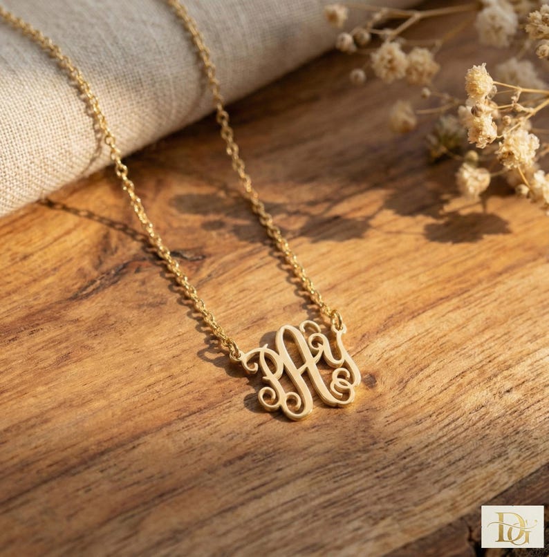 May include: A gold-colored monogram necklace with a delicate chain. The monogram features an ornate, intertwined design. The necklace is displayed on a wooden surface, with soft lighting and floral accents in the background.