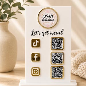 May include: A white rectangular acrylic sign featuring a gold-rimmed circular logo for K&D Antojitos Houston, TX. The text Let's get social is printed in black. It includes gold-bordered icons for TikTok, Facebook, and Instagram with QR codes.