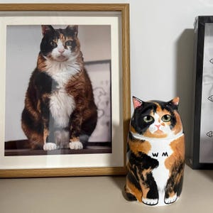 May include: A framed photograph of a calico cat next to a ceramic cat-shaped container. The container is black, orange, and white with the initials "WM" on its chest. The photograph and container share similar color patterns.