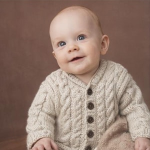 May include: A smiling baby wearing a beige cable-knit cardigan with dark brown buttons. The sweater has a classic design with a textured pattern. The baby has blue eyes and fair skin.