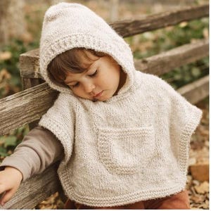 May include: A cream-colored knitted hooded poncho with a front pocket. The poncho is worn by a child, who is leaning against a wooden fence. The poncho is made of a textured yarn, and the hood is up. The child is wearing a long-sleeved shirt.