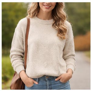 May include: A beige crew neck sweater with long sleeves. The sweater is paired with blue jeans. A brown leather shoulder bag is worn over the shoulder. The person is smiling, with hands in the pockets.