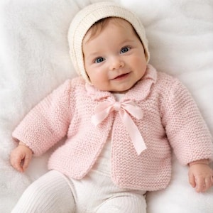 Puede incluir: Un bebé con una rebeca de punto rosa claro con un lazo a juego y un gorro color crema. El bebé también lleva mallas blancas. El bebé sonríe y mira a la cámara.