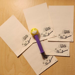May include: A collection of white notepaper with a black and white illustration of a car with a driver. A purple and yellow smiling face Pez dispenser is in the center of the image.