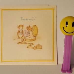 May include: A greeting card with a yellow border and the text "Time to say hi" featuring illustrations of mice and a pocket watch. A yellow smiley face PEZ dispenser with a purple base is to the right.