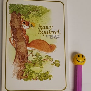 May include: A stationery gift set titled "Saucy Squirrel" featuring a cartoon squirrel illustration. The set is next to a purple and yellow smiley face Pez dispenser.
