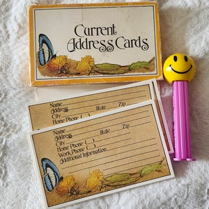May include: A vintage address card set with a yellow and orange box, featuring the text "Current Address Cards." The cards have a butterfly and floral design. A yellow smiley face PEZ dispenser is also present.