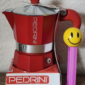 May include: A red Pedrini stovetop espresso maker with a silver top and handle. The espresso maker is next to a yellow smiley face Pez dispenser with a pink base. The Pedrini box is visible at the bottom.