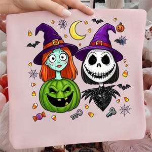 May include: A light pink fabric square featuring a cartoon illustration of two characters in purple witch hats. One character has orange hair and a green pumpkin body, while the other is a skeleton figure. Includes bats, spiderwebs, and candy corn.