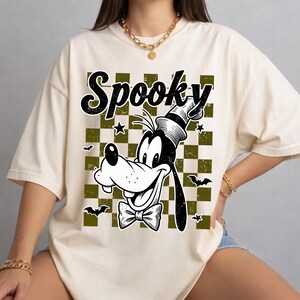 Könnte beinhalten: Ein cremefarbenes Kurzarm-T-Shirt mit einem Grafikdruck eines Cartoon-Hundes, der einen Zylinder und eine Fliege trägt. Das Design zeigt das Wort Spooky in schwarzer Schrift, einen olivgrünen und cremefarbenen Schachbrett-Hintergrund sowie kleine Fledermäuse.