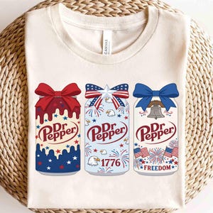 Pode incluir: Camiseta creme com três designs de latas de refrigerante Dr. Pepper com temas patrióticos. Cada lata tem um laço no topo e é decorada com estrelas vermelhas, brancas e azuis, fogos de artifício e as palavras "Dr. Pepper". Uma lata diz "1776" e outra "Freedom".