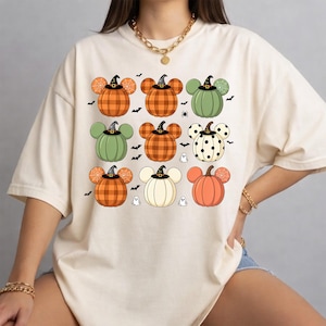 May include: A cream-colored short-sleeve t-shirt featuring a graphic print of nine pumpkin-shaped characters with mouse-like ears. The pumpkins display various patterns including plaid, polka dots, and spiderwebs, each wearing a small black witch hat.