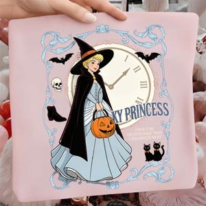 May include: A light pink fabric item featuring a cartoon illustration of a blonde character in a witch hat and black cape holding a pumpkin bucket. The design includes a clock face, bats, a skull, a boot, two black cats, and the text SPOOKY PRINCESS.