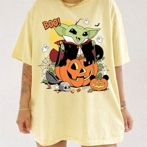 May include: A pale yellow t-shirt featuring a graphic of a green creature dressed as a vampire sitting on a large orange jack-o'-lantern. The design includes the text BOO!, small ghosts, a skull, candy corn, and a spider web against a yellow moon.