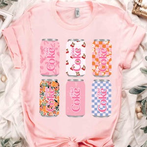 Può includere: T-shirt rosa chiaro con sei design di lattine Diet Coke. Le lattine presentano vari motivi, tra cui floreali, ciliegie e a scacchi, in tonalità rosa, arancione e blu. Il testo "Diet Coke" è visibile su ogni lattina.