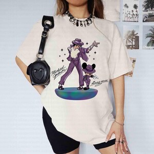 May include: Cream-colored t-shirt featuring a graphic of Michael Jackson and Mickey Mouse in matching purple outfits. The text "Michael Jackson" and "Mickey Mouse" are also on the shirt. The design is on a round, colorful stage.