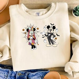Peut inclure: Un sweat-shirt à col rond couleur crème avec une impression graphique de deux personnages de dessin animé. L'un porte une robe en patchwork devant une toile d'araignée, l'autre un costume rayé. Des fantômes, chauves-souris et citrouilles entourent le tout.