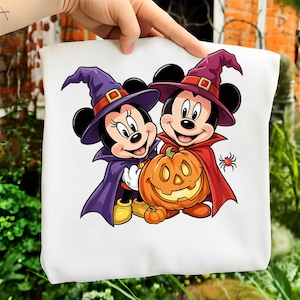 May include: A white t-shirt featuring a graphic of two cartoon mice dressed as witches, wearing purple hats and capes, standing next to a carved orange jack-o'-lantern pumpkin with a small spider nearby on a light background.