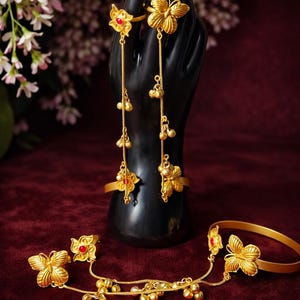 May include: Gold-colored jewelry set featuring butterfly and floral designs with red accents. The set includes rings, a bracelet, and a hand chain, all displayed on a black mannequin hand against a burgundy background.
