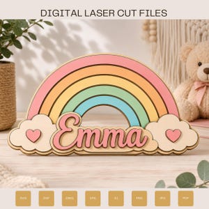 Op de afbeelding: Een houten regenboogvormig bord met de naam Emma in roze cursief schrift. De regenboog heeft pastelkleurige bogen in roze, geel, groen en blauw. Twee wolken met kleine roze hartjes bevinden zich aan de zijkanten. Tekst luidt DIGITAL LASER CUT FILES.