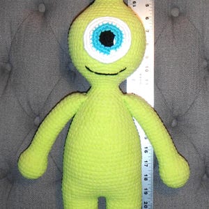 Puede incluir: Un peluche de ganchillo verde lima con forma de monstruo de un ojo. El ojo es blanco, azul y negro. El juguete mide aproximadamente 23 cm de alto. Tiene cuello largo, brazos y piernas. El juguete está sobre un fondo gris.