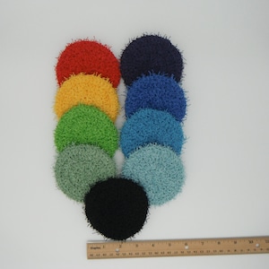 Puede incluir: Una colección de diez estropajos redondos de ganchillo en varios colores, incluyendo rojo, amarillo, verde, azul y negro. Una regla de madera en la parte inferior muestra las medidas en pulgadas (aproximadamente 30 cm).