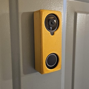 Może przedstawiać: Żółta prostokątna kamera dzwonka zamontowana na szarych drzwiach. Kamera ma okrągły obiektyw na górze i czarny przycisk na dole. Dzwonek jest umieszczony w żółtej ramie.