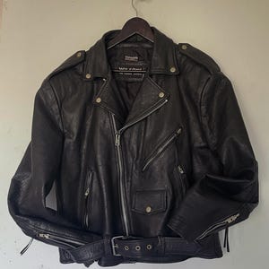 May include: Black leather biker jacket with a notched collar, multiple zipped pockets, and a belt at the waist. The jacket has silver hardware and a classic biker style. The jacket is hanging on a wooden hanger.