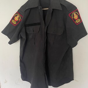 Puede incluir: Una camisa negra de manga corta con dos bolsillos en el pecho y cuello. Parches en ambas mangas con la inscripción "Canadian Forces, Military Police, Gendarmerie". La camisa está colgada en una percha de madera.