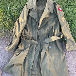 May include: An olive green military trench coat with a double-breasted design, belt, and epaulets. The coat features gold chevrons and a red patch on the sleeves. The coat is lying on a grassy surface.