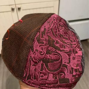 May include: A brown tweed flat cap with a burgundy embroidered design. The cap features a detailed pattern on the side and a small orange accent. The cap is a classic style, suitable for various occasions.
