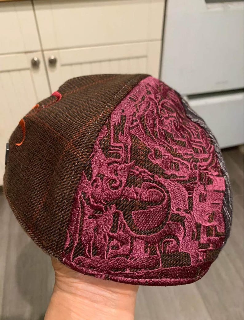 May include: A brown tweed flat cap with a burgundy embroidered design. The cap features a detailed pattern on the side and a small orange accent. The cap is a classic style, suitable for various occasions.