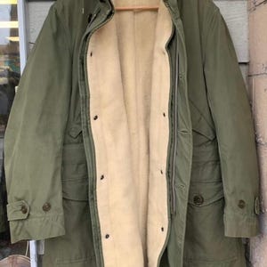 Puede incluir: Un abrigo de invierno verde oliva con capucha y forro afelpado color crema. El abrigo tiene cierre de cremallera, detalles de botones y bolsillos. El abrigo está colgado de una percha de madera.