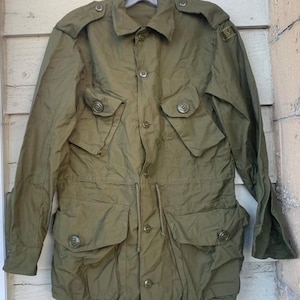 Op de afbeelding: Olijfgroene jas in militaire stijl met meerdere zakken en knoopsluitingen. De jas heeft een taille met trekkoord en epauletten op de schouders. Een klein embleem is zichtbaar op de bovenarm. De jas hangt aan een hanger.